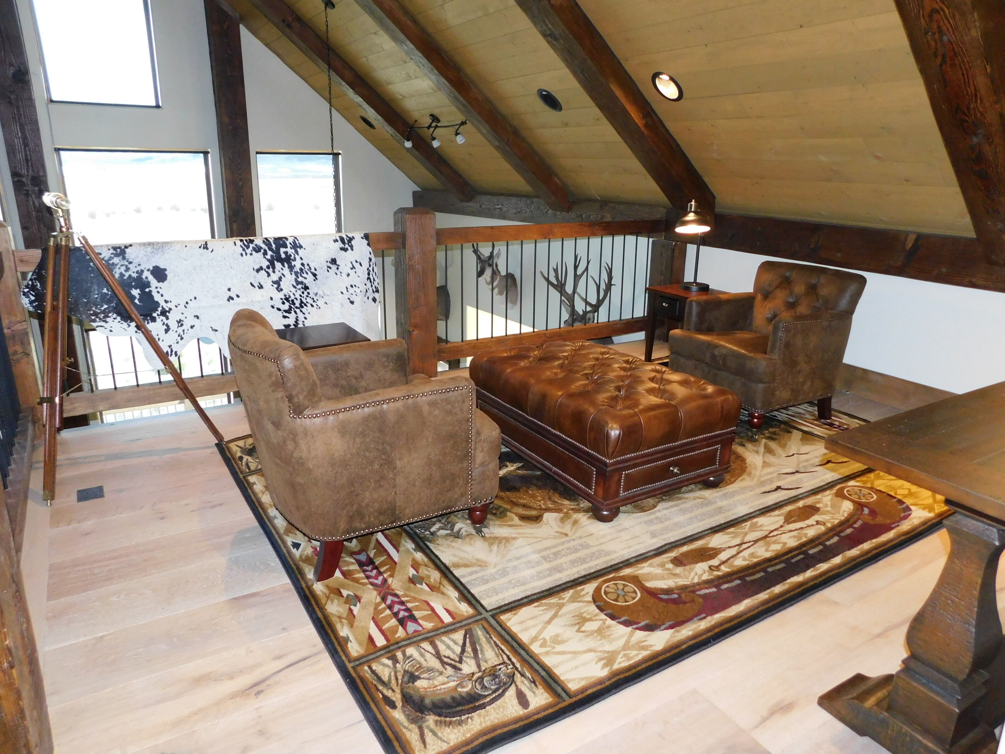Il s'agit d'une pièce de style « loft », avec un plafond haut, des poutres apparentes en bois et une grande fenêtre. On y trouve un canapé en cuir marron, deux fauteuils et une table basse en bois. Le sol est recouvert d'un tapis à motifs.