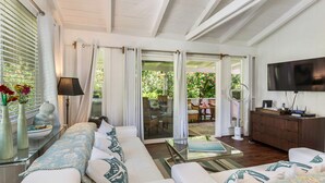 Ein geräumiges Wohnzimmer mit einer Couch, einem Glastisch und einem Fernseher. Eine Glastür führt zu einem Außenbereich mit Esstisch und Stühlen.