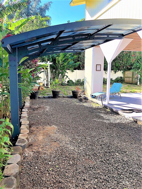 A covered patio area with a gravel ground, surrounded by potted plants and a blue chair. - Gated cottage, 3 blocks to beach, A/C Close to Tunnels. Lots of outdoor space (Haena)