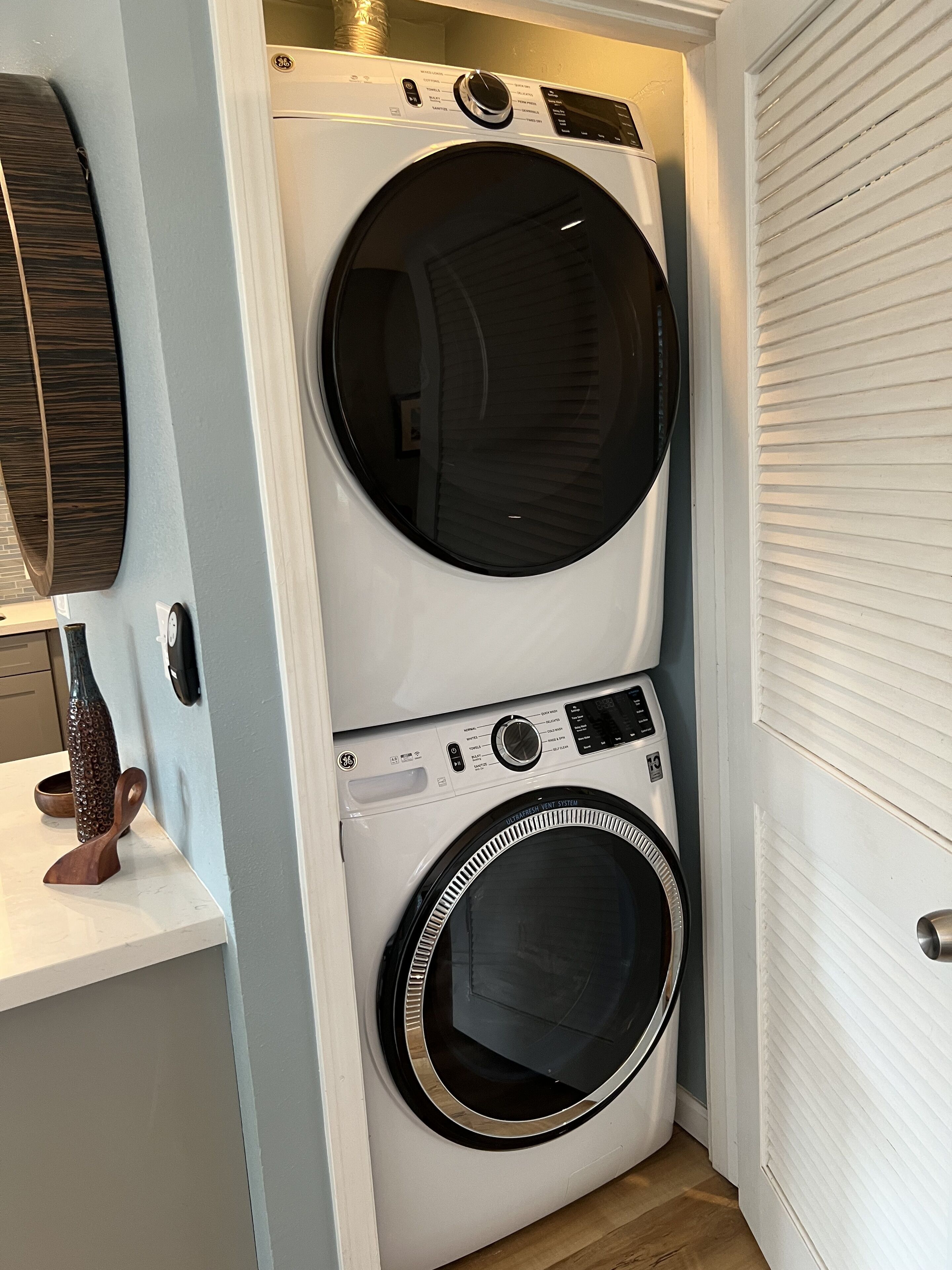A compact laundry room with a stacked washer and dryer, a wooden wall clock, and a small wooden sculpture on a white countertop.