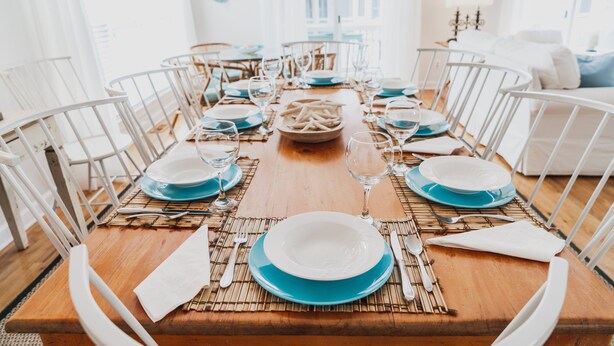 Una mesa de comedor dispuesta con platos azules y blancos, cubiertos y copas, rodeada de sillas blancas.