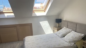 A bedroom with a skylight, a bed with floral bedding, a bedside table with a lamp, and a view of buildings through the skylight. - Studio in Paris, Le Marais (Paris)