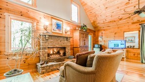 A cozy cabin interior with a stone fireplace, wooden walls, a comfortable sofa, and a television. - ♨️Hot Tub🛏️King Bed🔥Fireplace 🍷Wineries🌲Oasis🥾Hike🌎Comfy🔥Fire Pit⛲Pond 🆒 (Dahlonega)