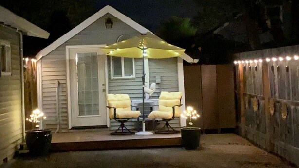A small, single-story house with a porch featuring two chairs and a table, surrounded by a fence.