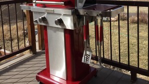 Een rode buitenbarbecue op een houten terras, met uitzicht op bergen en een rustiek gebouw op de achtergrond.