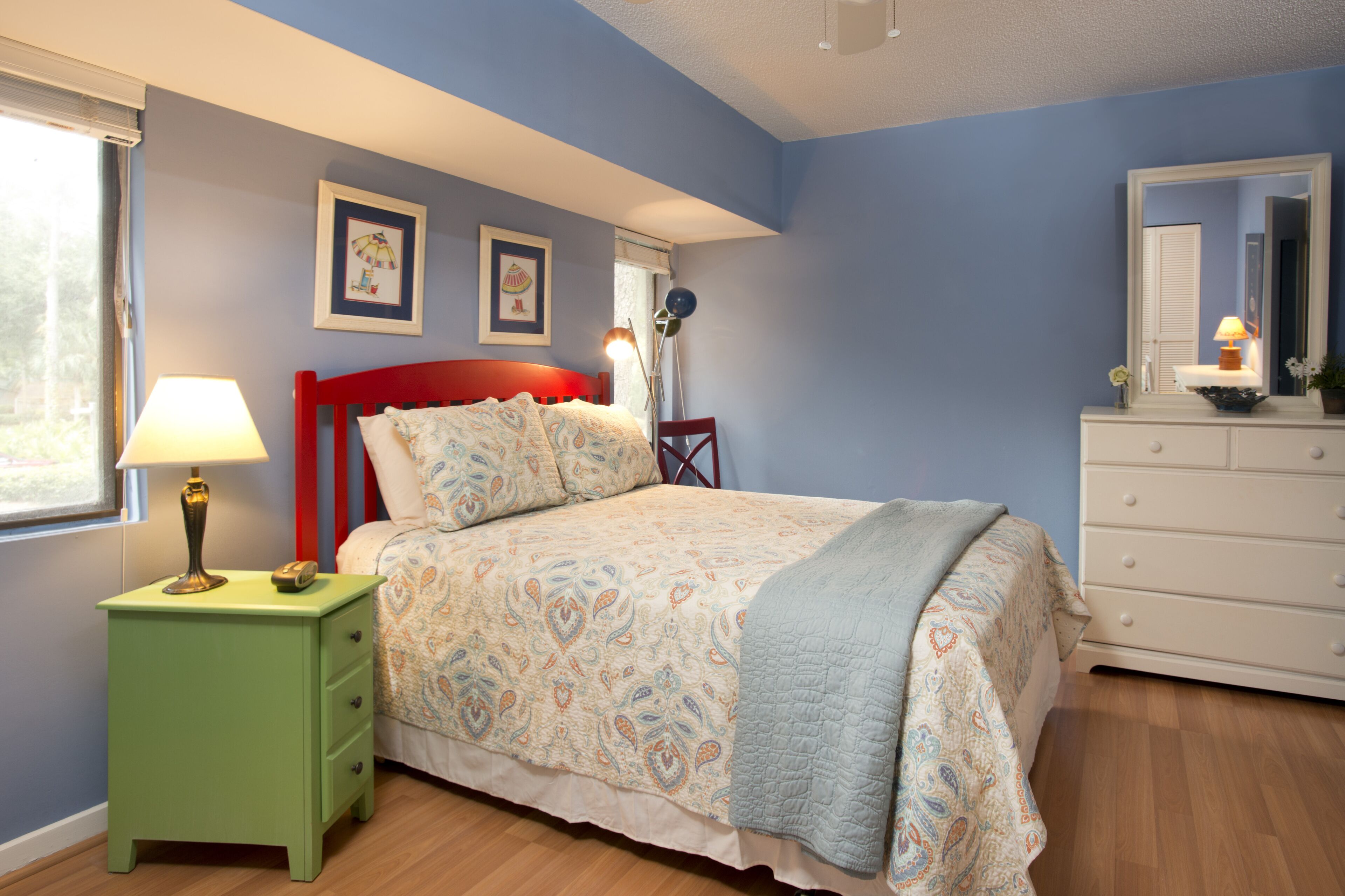 Une chambre à coucher comprenant un lit, une table de chevet, une commode, une fenêtre et deux tableaux accrochés au mur.