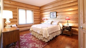 A cozy log cabin bedroom with a wooden bed, two bedside lamps, a window, and a framed picture on the wall. - Secluded Log Home with Mountain View (Granby)