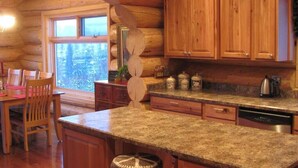 A log cabin kitchen with wooden cabinets, granite countertops, and a dining area with chairs. - Secluded Log Home with Mountain View (Granby)