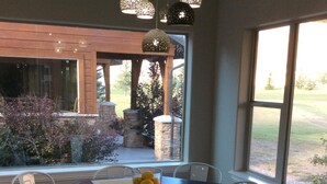 A dining area with a dark wooden table, white chairs, and a bowl of lemons. There are hanging lights and a view of a garden outside.