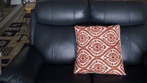 A modern living room with a black leather sofa, a patterned throw pillow, and a glass-top coffee table. In the background, there is a kitchen with white cabinets, stainless steel appliances, and a dining area with a round table and chairs. - Beautiful Oceanfront  Condo- Crescent Shores - Privately Owned and Maintained (North Myrtle Beach)