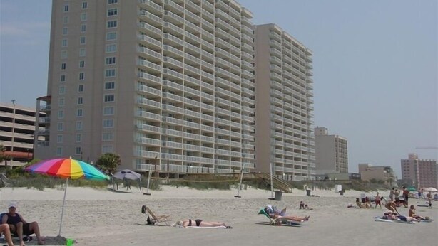 A high-rise hotel with multiple balconies, located near a beach with people sunbathing and a colorful umbrella. - Beautiful Oceanfront  Condo- Crescent Shores - Privately Owned and Maintained (North Myrtle Beach)