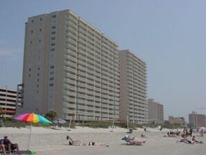 A high-rise hotel with multiple balconies, located near a beach with people sunbathing and a colorful umbrella. - Beautiful Oceanfront  Condo- Crescent Shores - Privately Owned and Maintained (North Myrtle Beach)