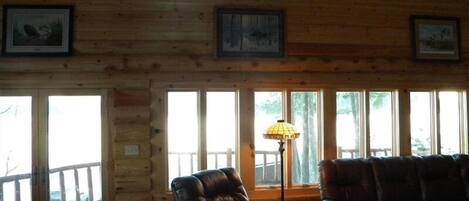 A log cabin interior with a leather armchair and sofa, large windows, and framed artwork on the walls.