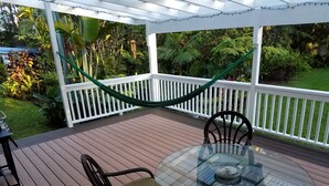 Eine überdachte Terrasse im Freien mit einem Glastisch und Stühlen, umgeben von einem weißen Zaun und üppigem Grün.