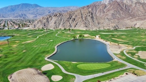 Un campo de golf con un estanque, rodeado de montañas y zonas residenciales.