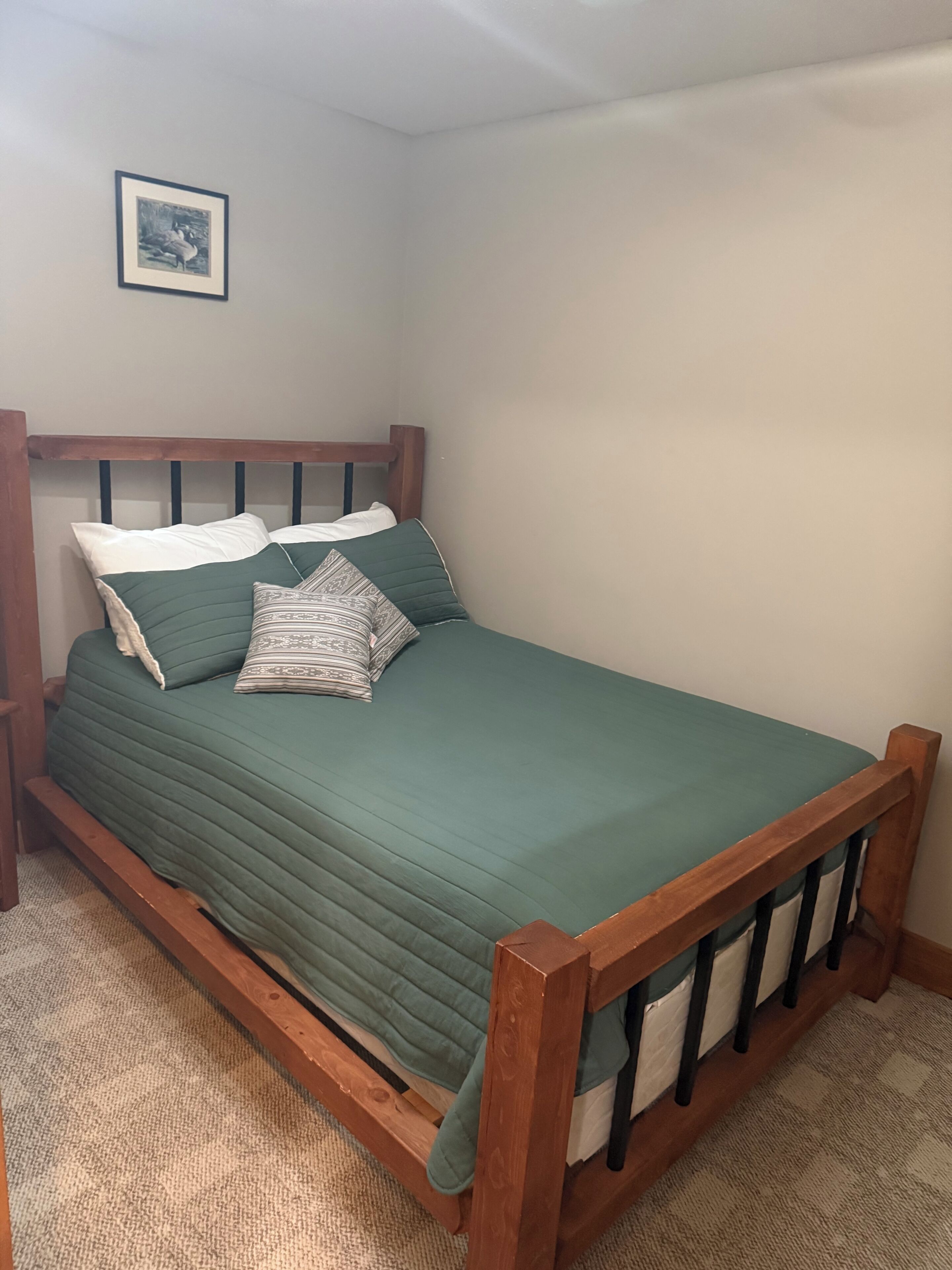 Una cama individual con estructura de madera y colcha verde, acompañada por dos almohadas y un cojín decorativo.