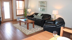 A living room with a black leather sofa, two wooden coffee tables, a patterned rug, and a glass door leading outside. - Experience a great family vacation at Casa Del Mila Oro ! (Osoyoos)