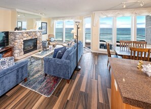 Een ruime woonkamer met een stenen open haard, een grote bank, een eethoek met uitzicht op de oceaan en een balkon met buitenmeubels.
