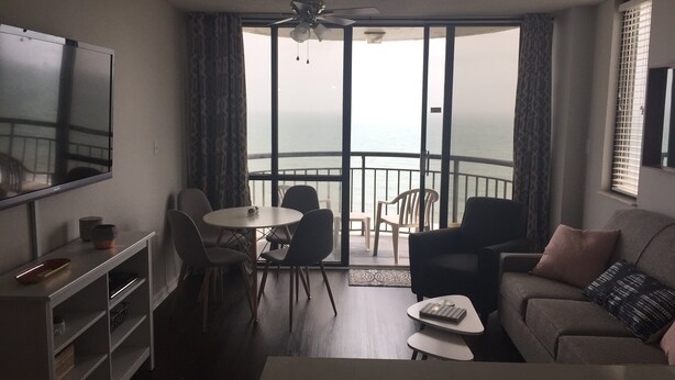A living room with a sofa, a coffee table, and a TV on a stand. There is a balcony with a view of the sea. - Brand New Reno! OF, End Unit, King bed, Quartz Counters, Shaker Cabinets (Myrtle Beach)
