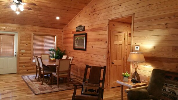 Interior acogedor de una cabaña de madera, con comedor, una puerta que dice 'WELCOME' y una pequeña mesa con una lámpara.