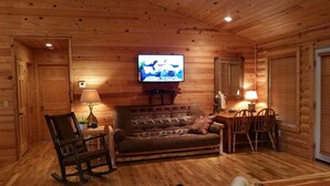 Acogedor interior de cabaña de madera con una televisión sobre un soporte, una mecedora, un sofá y un comedor con mesa y sillas.
