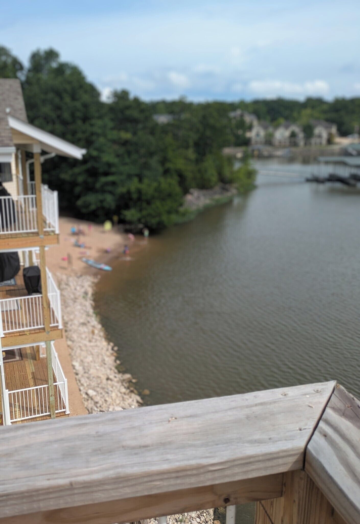 A river with a wooden deck overlooking it, a beach with people and boats, and a building with balconies.