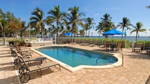 Ein Poolbereich mit Liegestühlen und Sonnenschirmen, umgeben von Palmen und Blick auf das Meer.