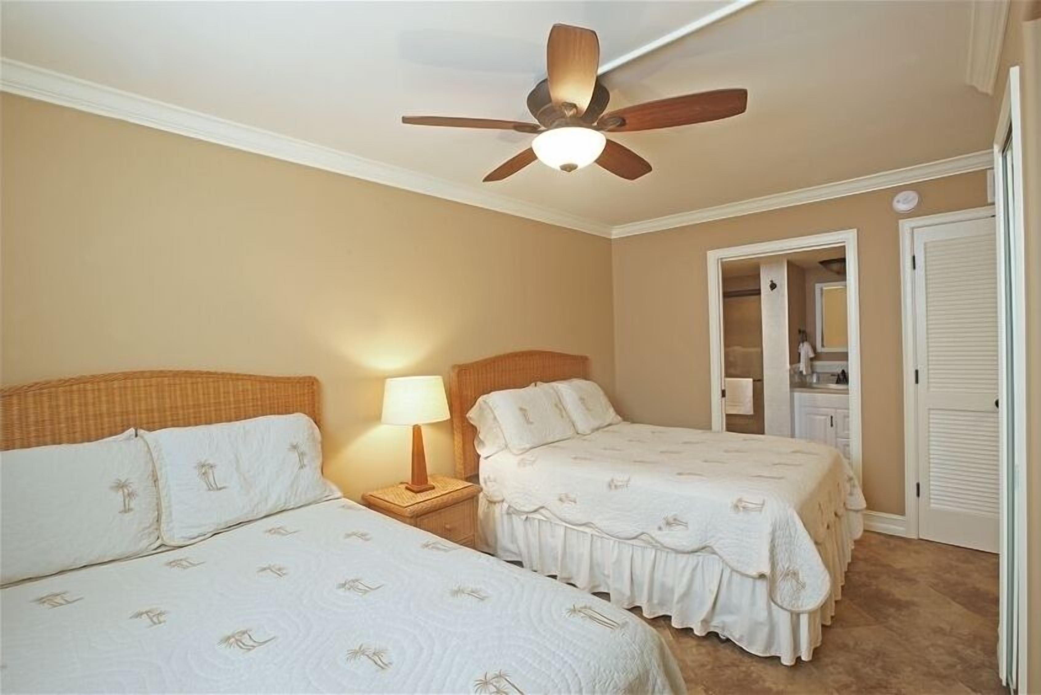 A bedroom with two beds, a ceiling fan, a nightstand with a lamp, and a bathroom visible through an open door.