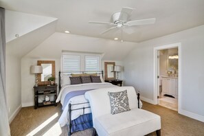 Une chambre à coucher comprenant un lit, des tables de chevet, une chaise, un ventilateur de plafond et une salle de bain visible à travers une porte ouverte.