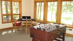 A room with a foosball table, a dining table with chairs, and large windows with a view of trees. - Golf Course/Lodge Style/large/clean/comfortable/5 BR (Black Butte Ranch)