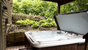Een hottub in een openlucht setting met een houten terras, omgeven door groen en een stenen muur.