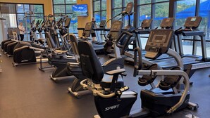 Een sportschool met loopbanden en hometrainers, grote ramen en een televisiescherm.