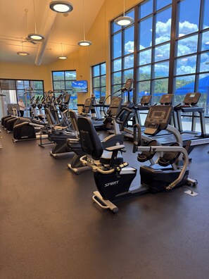 Un gimnasio con cintas de correr y bicicletas estáticas, ventanas amplias y una pantalla de televisión.