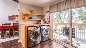 A laundry area with two front-loading washing machines, a wooden table with chairs, a shelf with books and cleaning supplies, and a view of the outdoors through a sliding glass door. - Spacious Beach House (Solana Beach)