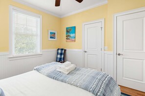 A bedroom with a bed, a window with blinds, a door, and two paintings on the wall.