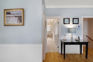 A hallway with a framed landscape painting, a black table with a lamp, and a door leading to a bathroom.