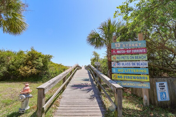Un muelle de madera que conduce a una playa con varios carteles que indican normas como prohibido animales, vidrio, basura, fuegos artificiales, y fumar o vapear.