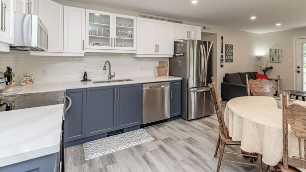 A modern kitchen with white and blue cabinets, stainless steel appliances, and a dining area with a patterned rug and wooden chairs. - 4 Season Cottage at Lake Vernon - Perfect for families! (Huntsville)