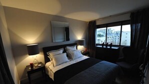 Een hotelkamer met een bed, twee bedlampen, een nachtkastje, een stoel en een groot raam met uitzicht op gebouwen.