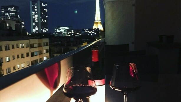 Een balkon met twee wijnkannen en een kaars, met uitzicht op een stadsbeeld met verlichte gebouwen en de Eiffeltoren op de achtergrond.