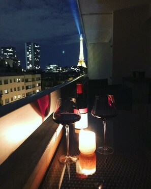 A balcony with two wine glasses and a candle, overlooking a cityscape with illuminated buildings and the Eiffel Tower in the background.