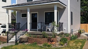 Ein zweigeschossiges Haus mit Veranda, überdachter Eingangspartei und einem kleinen Gartenbereich mit Pflanzen und einem Steinelement.