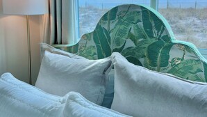 Ein Bett mit weißer Bettwäsche und einem Blattmuster an der Kopfteil, eine Lampe und Blick auf den Strand durch ein Fenster.