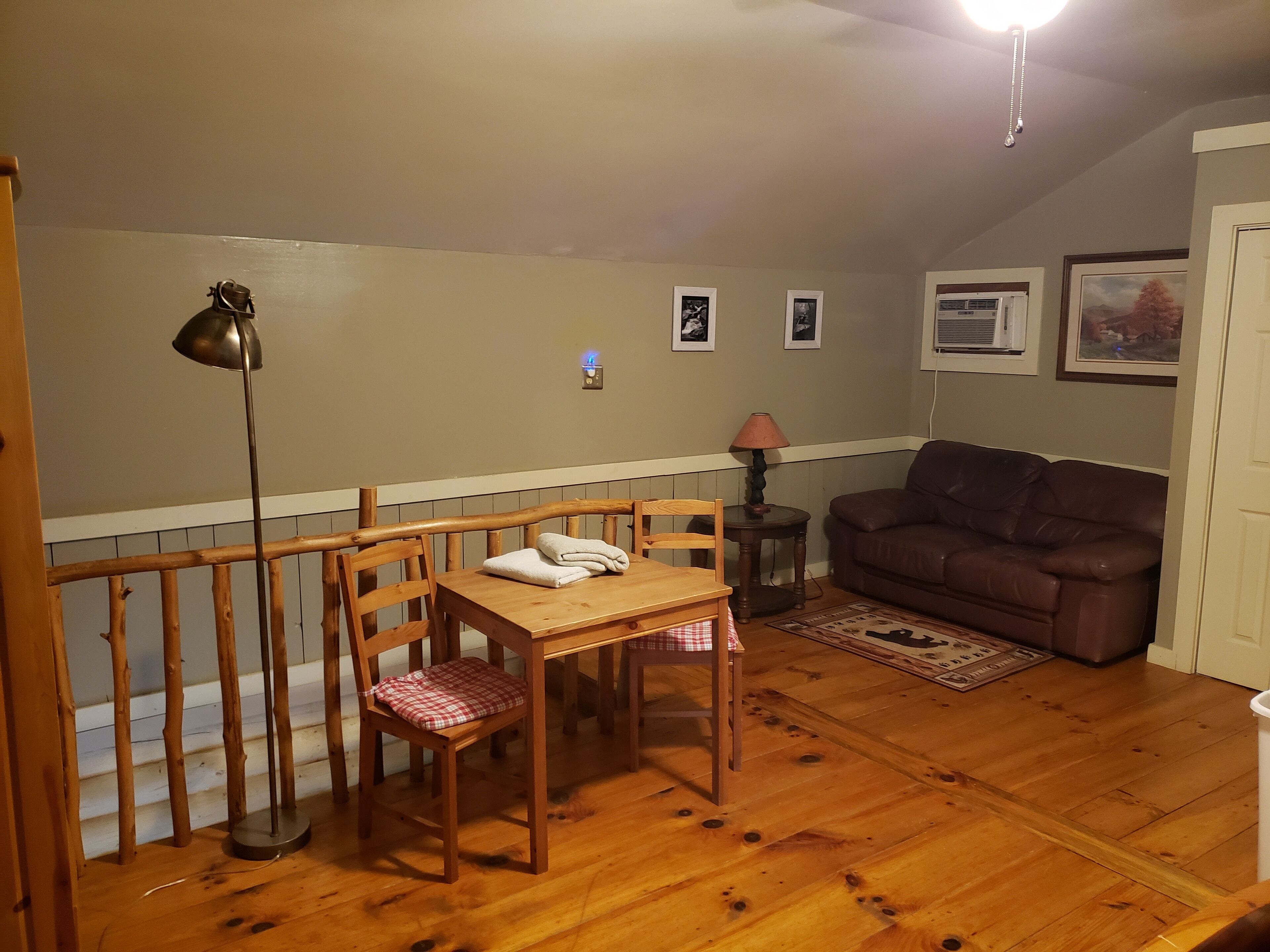 Een gezellige woonkamer met een houten eettafel, twee stoelen, een bruine leren bank, een vloerlamp en een klein vloerkleed.