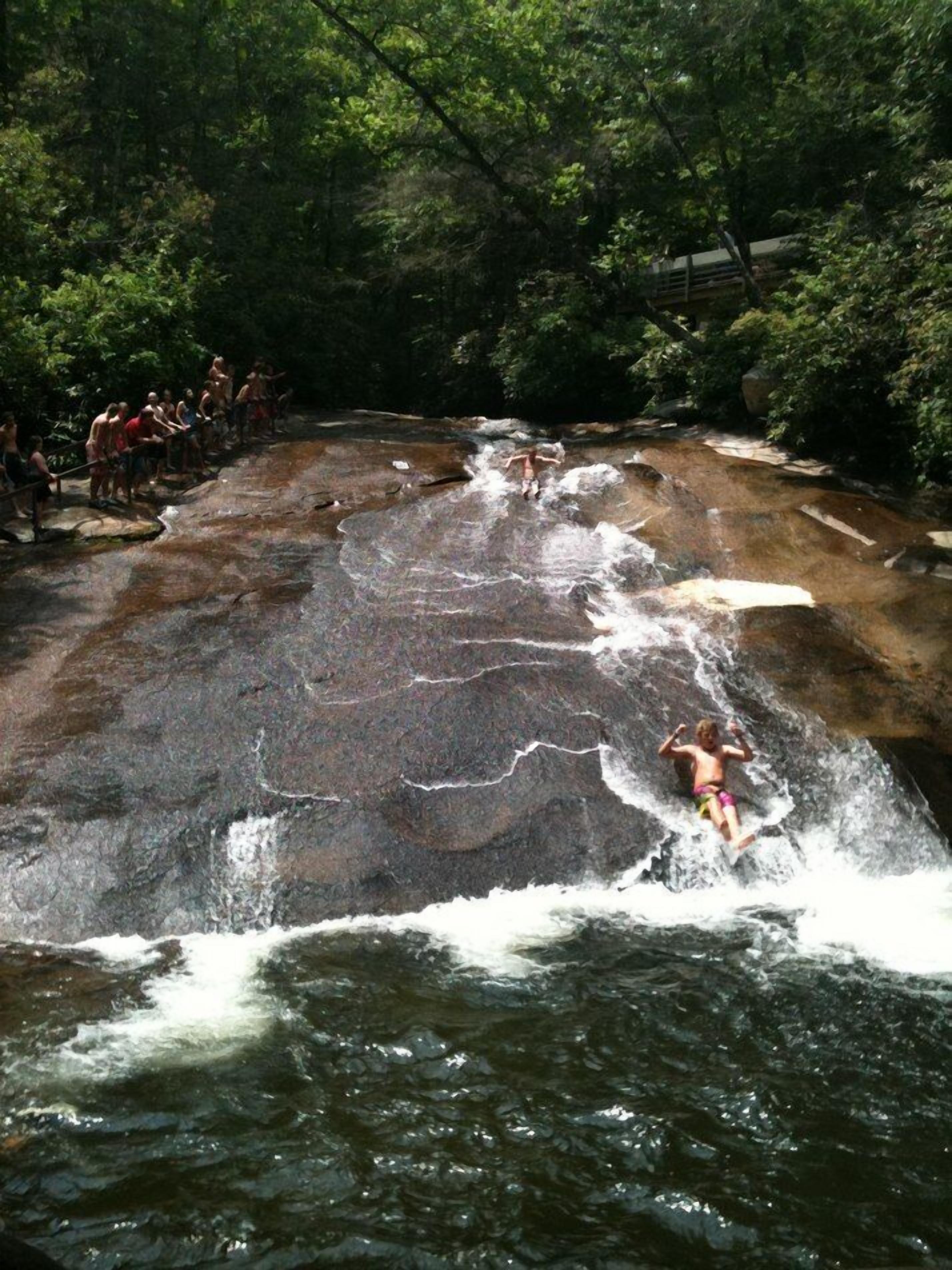Une cascade où l’on voit des personnes debout sur les rochers et une personne qui saute dans l’eau.