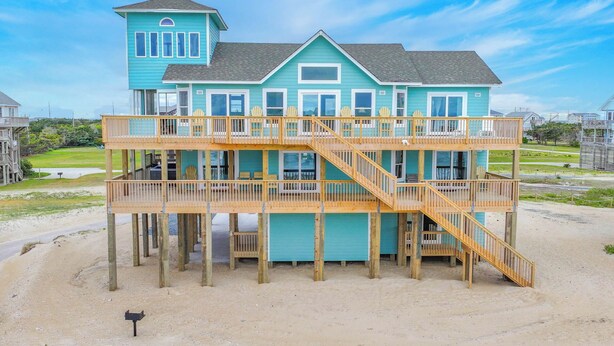 Ein zweistöckiges Strandhaus mit einer Holzterrasse und einer Treppe, die zum Obergeschoss führt.