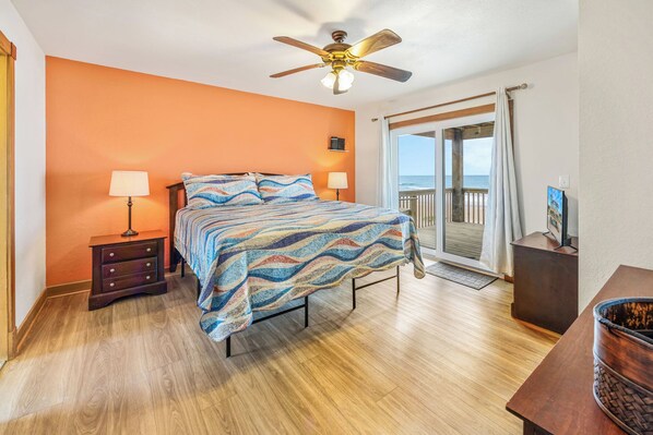 Un dormitorio con cama, dos mesitas de noche, ventilador de techo, vista al océano y piso de madera.