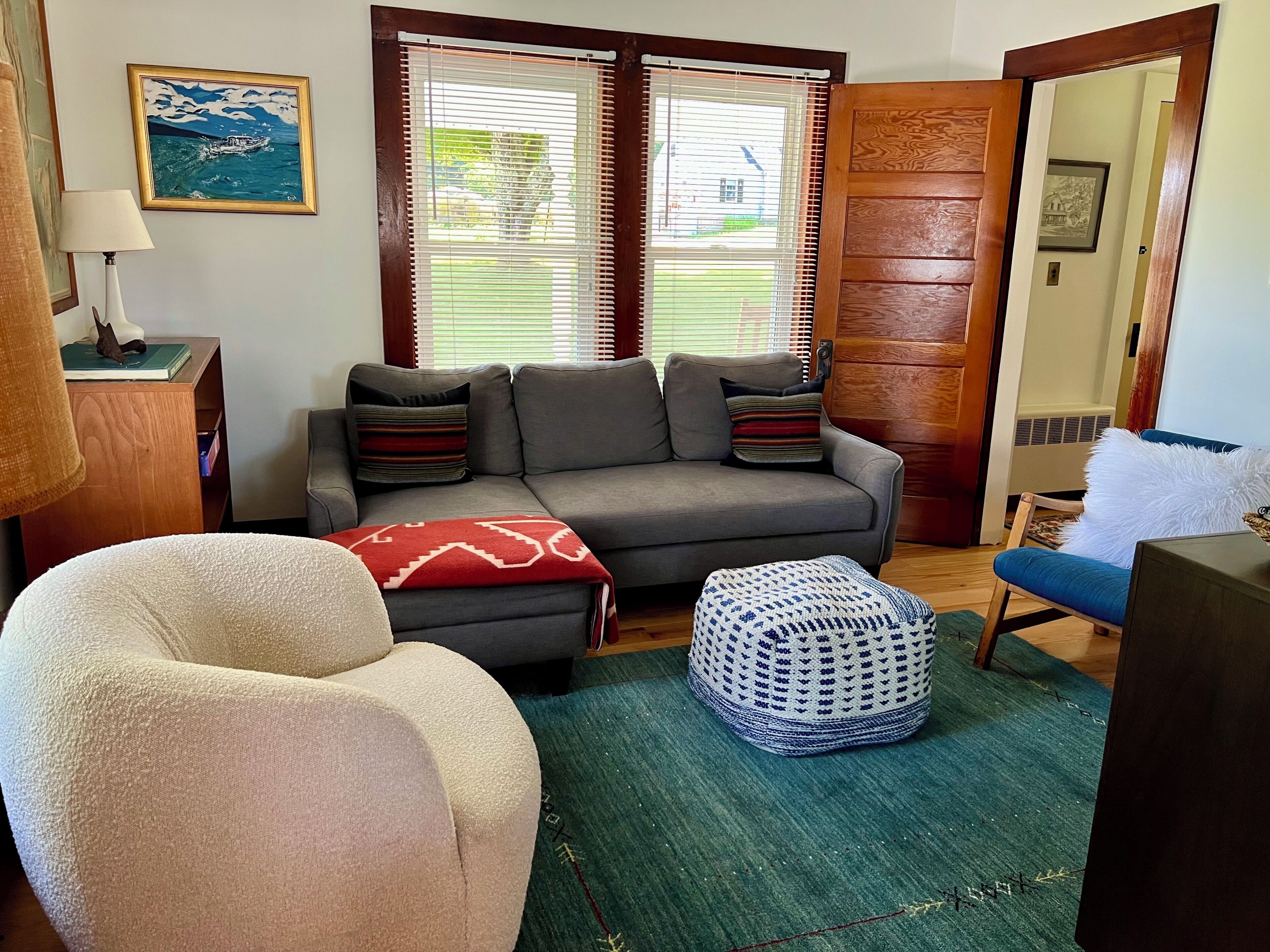 A living room with a grey sofa, a white armchair, a patterned ottoman, and a green rug.