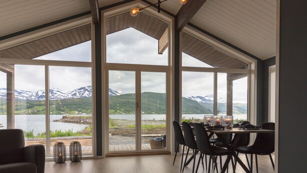 Un comedor con una mesa dispuesta para dos, amplias ventanas que ofrecen vistas a montañas y un lago, y un diseño de techo moderno.