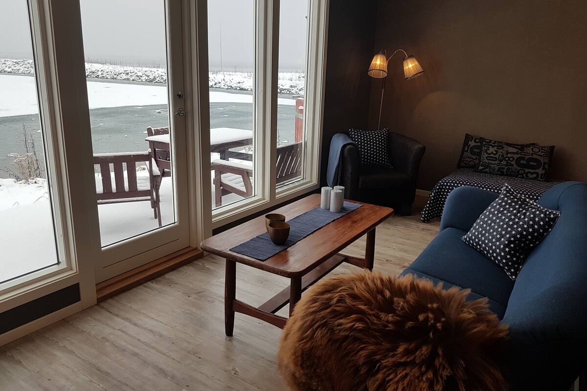 A cozy living room with a wooden coffee table, a blue sofa with patterned pillows, and a view of a snowy outdoor deck through large glass doors.